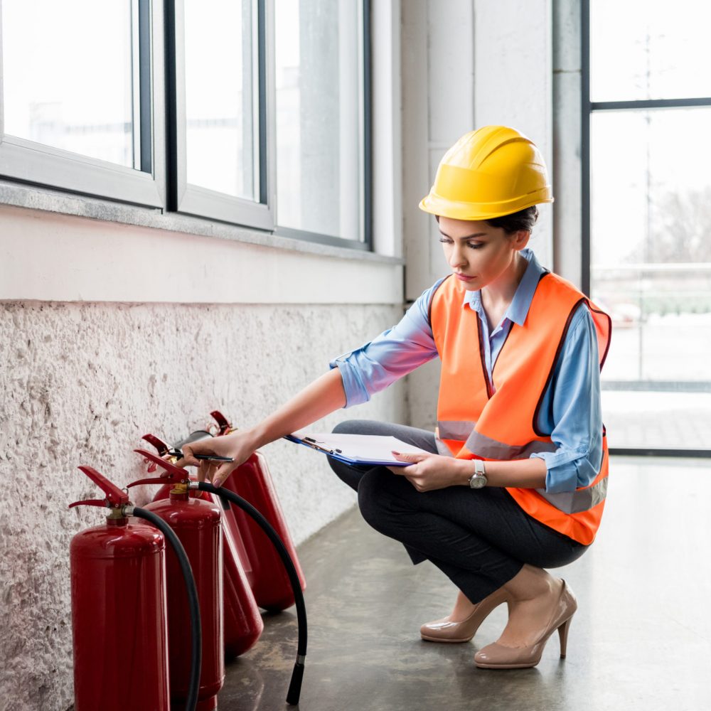 attractive firefighter in helmet holding clipboard and pen while checking extinguishers