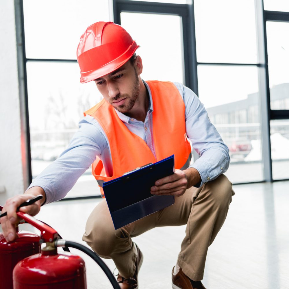 serious fireman in helmet checking extinguishers while sitting with clipboard